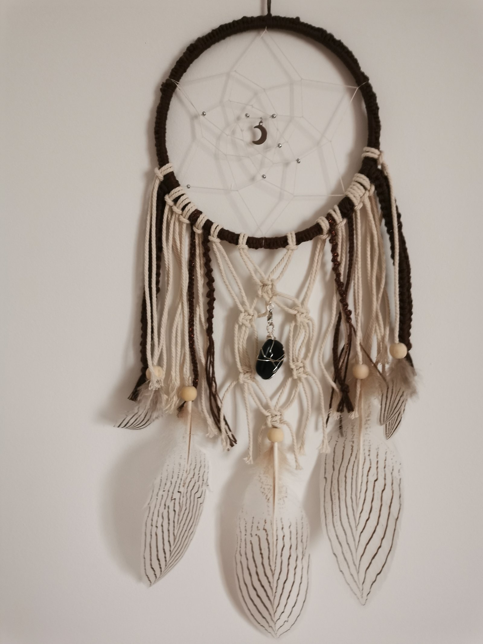Photographie d'un attrape-rêves artisanal Yudreami terminé, suspendu à un mur. Création en macramé avec des tissages complexes, des plumes tachetées suspendues et des détails de perles et cristaux.