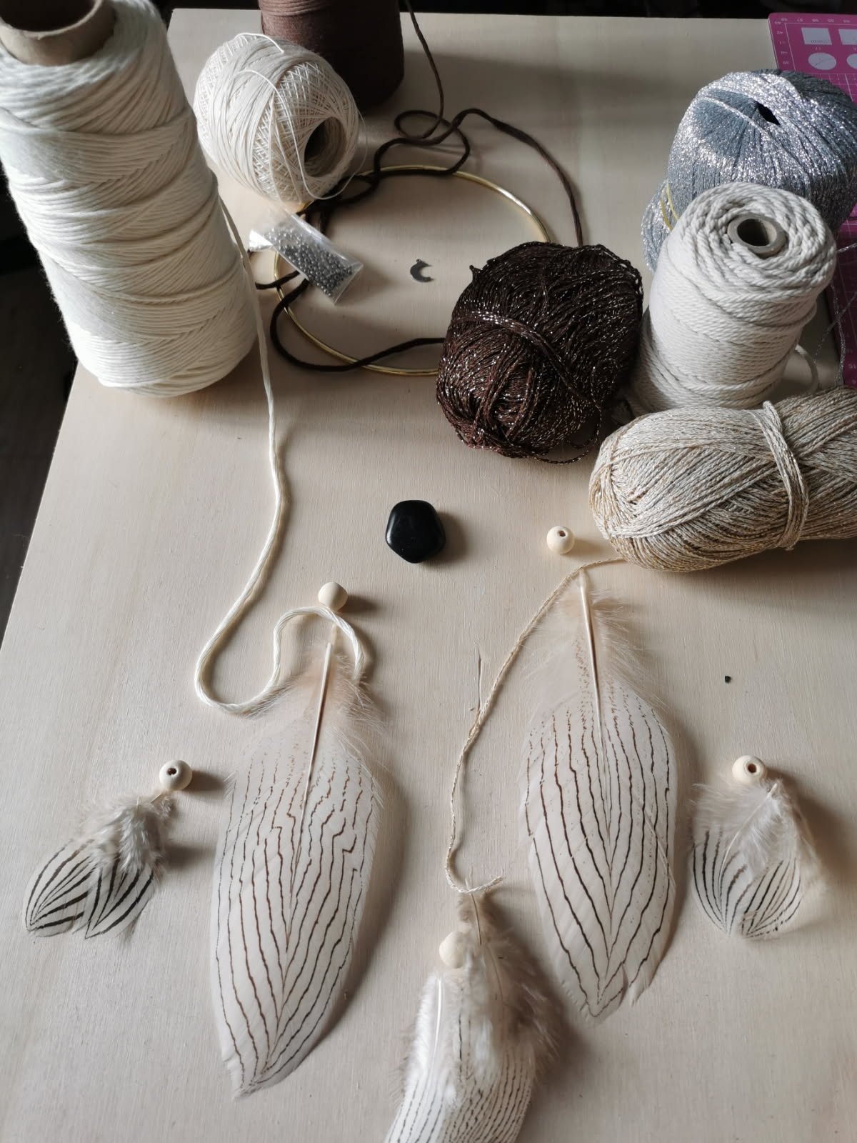 Vue de dessus de bobines de fil, plumes tachetées et matériel de création. Texte : Étape du Partage où nous validons ensemble le lancement de la création personnalisée. C'est à ce moment que je vous communique le prix final selon les matériaux sélectionnés.