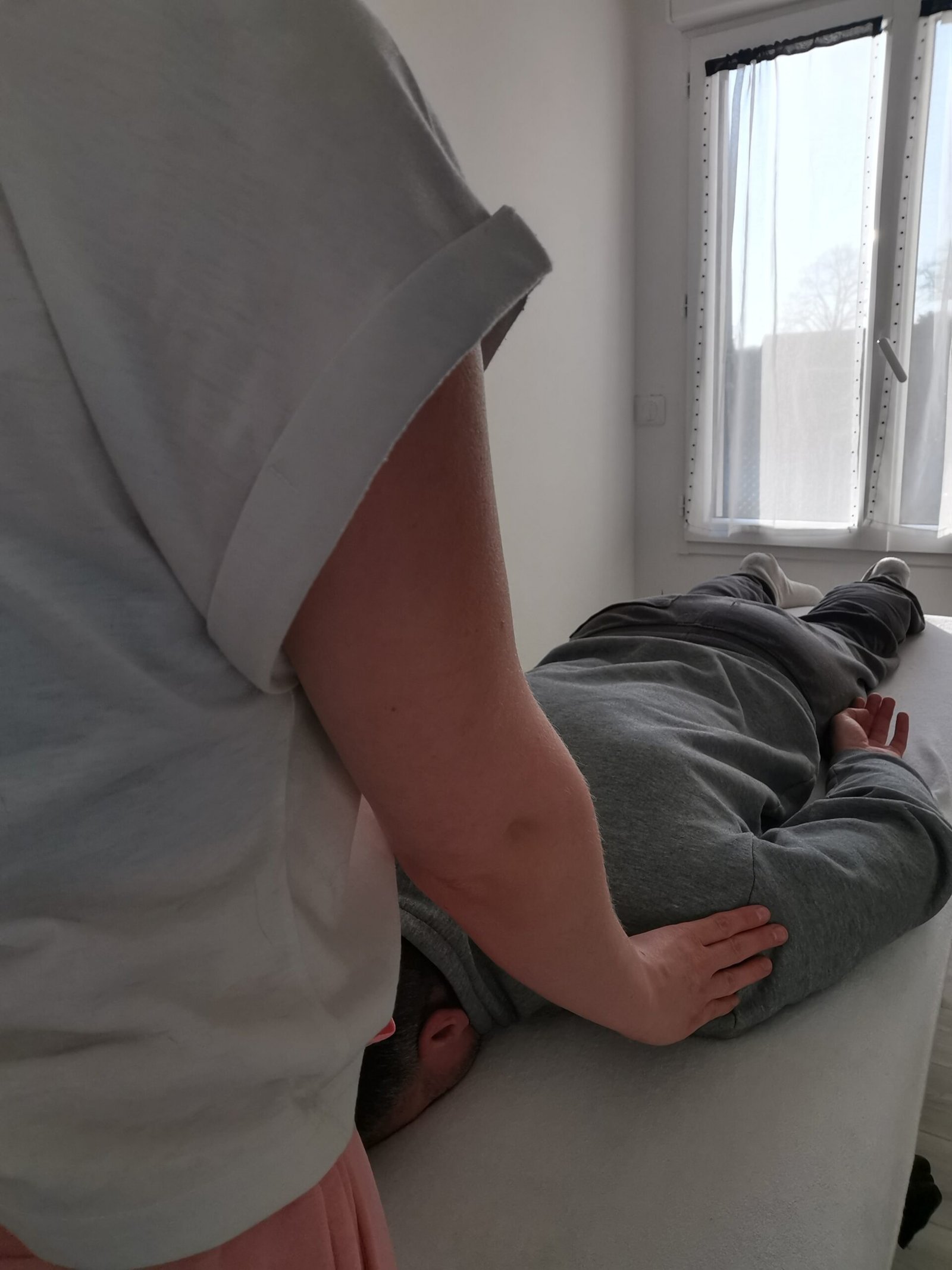 Vue de profil d'une séance de soin : Anaïs pose ses mains sur les épaules d'une personne allongée sur une table de massage.